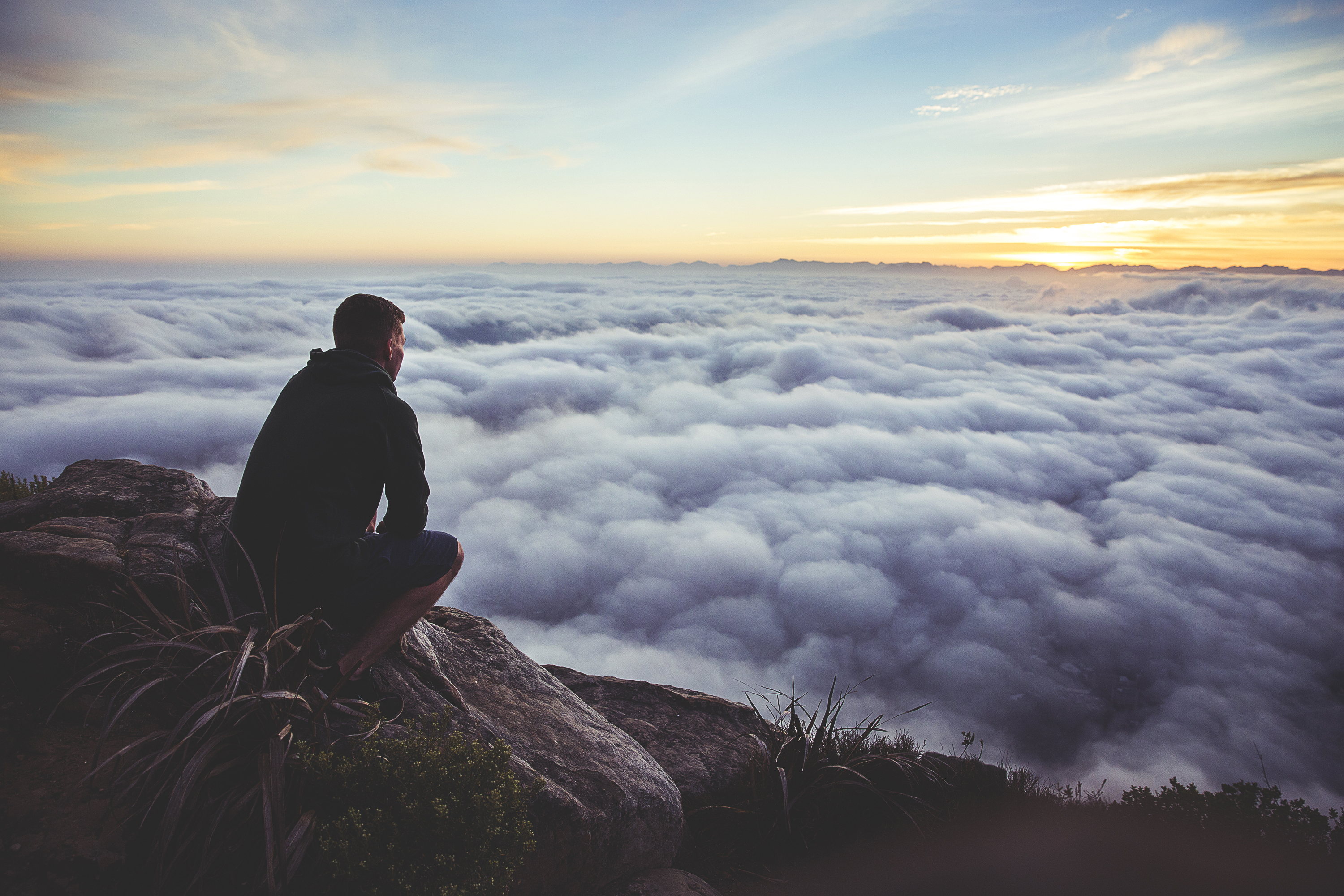 man crouching on a rock overlooking a the tops of clouds and a sunrise.