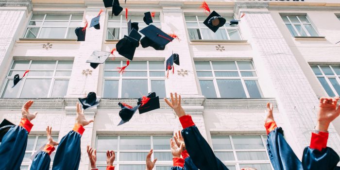 College graduates throwing their blue caps with red tassels into the air next to a tall building with windows.