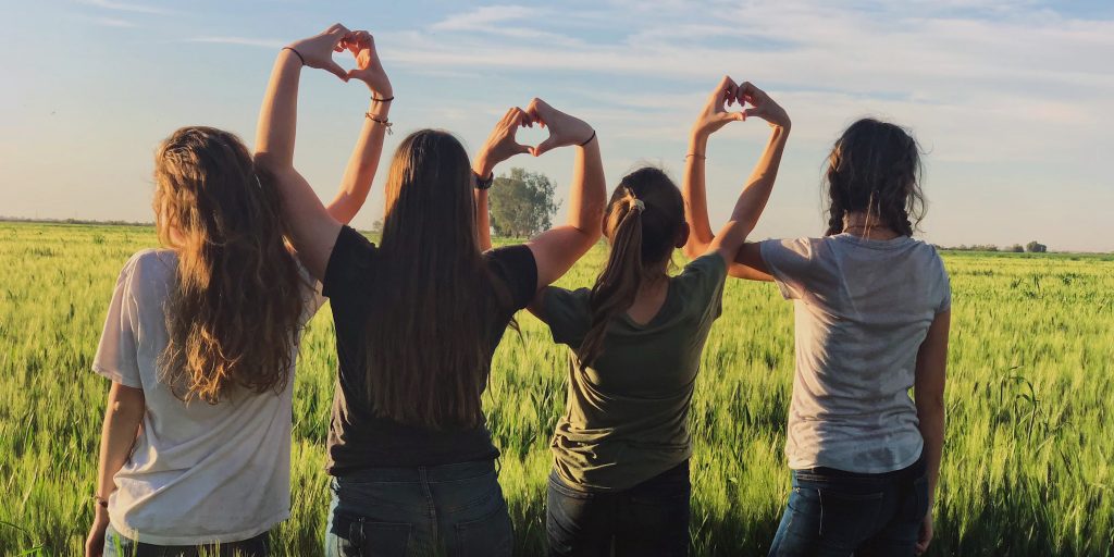 Four girls sitting in a grassy field, facing away from the camera, holding their hands together to form hearts