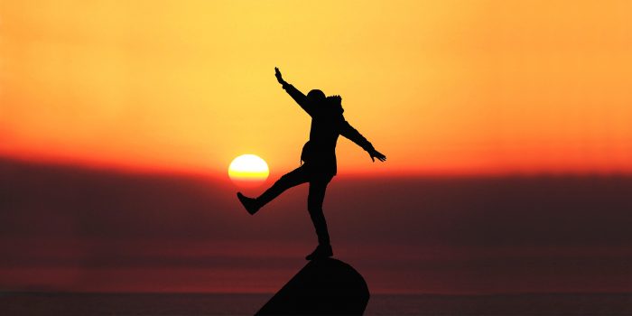 silhouette of a person balancing on a boulder on the beach, sun setting in the background.