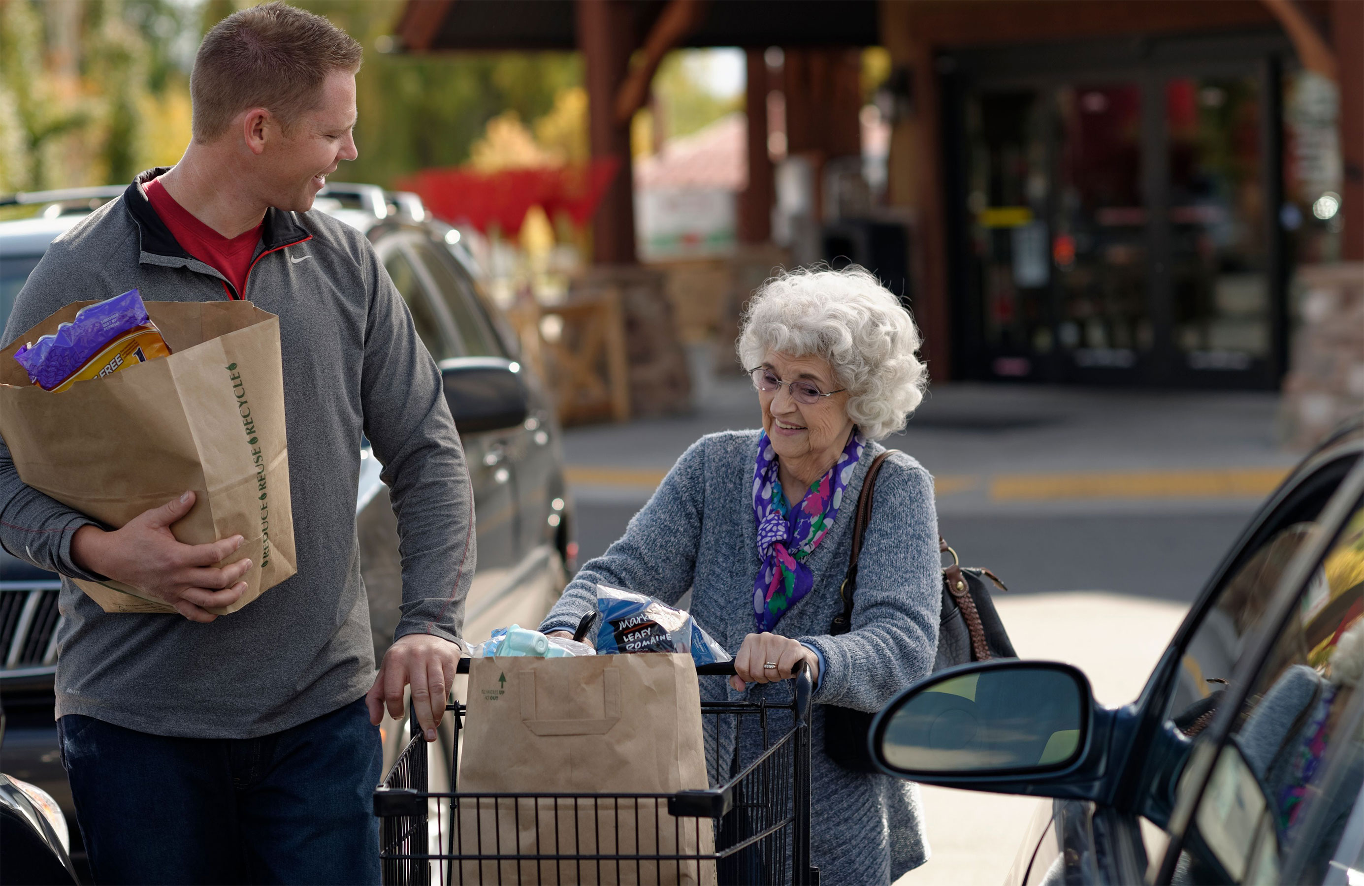 A young man helps an elderly woman carry groceries out to her car.