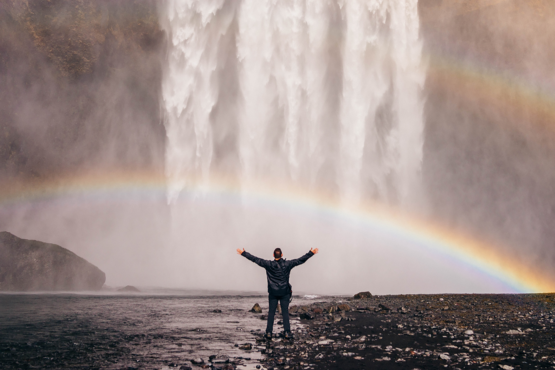 Silhouette of person with arms outstretched standing at the bottom of a waterfall and rainbow power couple