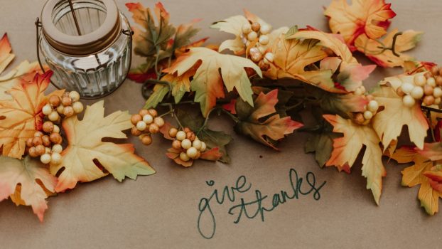 A surface is shown from above with fall leaf decorations, a mason jar, and the words “give thanks.”