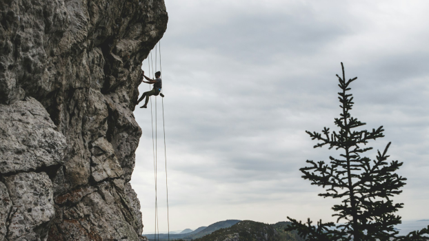 Person participating in outdoor rock climbing against a backdrop of cloudy skies, trees, and mountains.