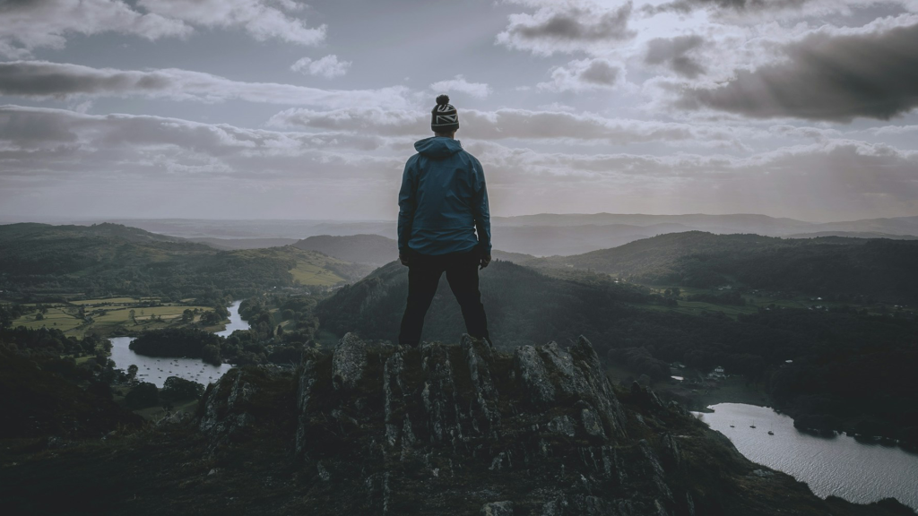 Person stands on a mountain and looks over more mountains with a river running through them