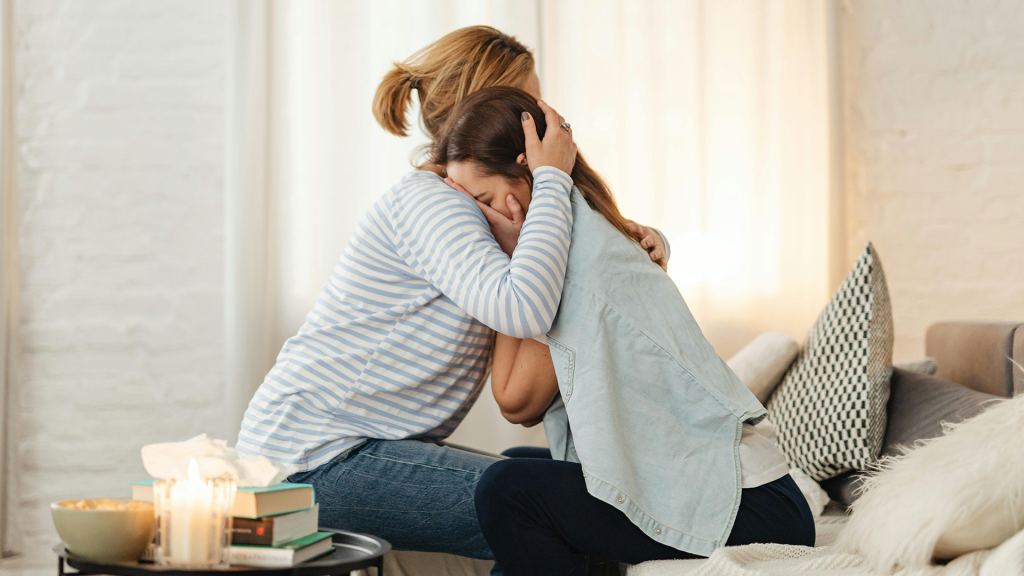 A woman in a striped shirt with short blond hair in a ponytail cradles the head of another woman with dark hair in comfort. They are both sitting on a bed.