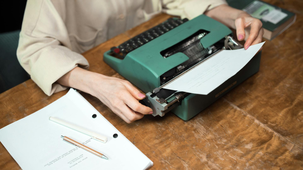 A woman sits at a desk and uses a green typewriter next to a manuscript and writing materials.